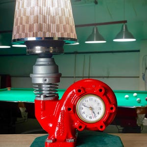 May include: A unique table lamp with a red, industrial-style base featuring a clock face. The lamp has a silver-colored body, a beige lampshade, and sits on a green, velvet-covered base. The clock shows the time.