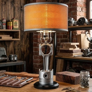 Puede incluir: Una lámpara de mesa única con una base de pistón metálico y una pantalla cilíndrica. La pantalla tiene un exterior de malla gris y un interior naranja. La lámpara se asienta sobre una base circular negra. El fondo incluye herramientas y estanterías de madera.