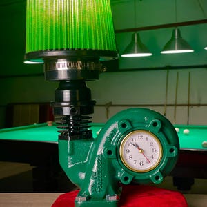 May include: A unique desk lamp featuring a green air filter shade, a black engine component body, and a green turbine housing with a clock face. The lamp sits on a red base, with a pool table in the background.
