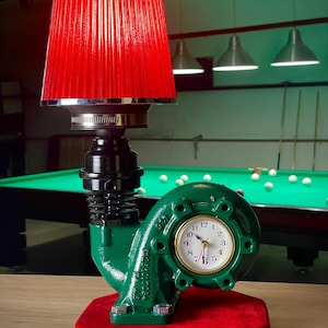 Op de afbeelding: Een unieke bureaulamp met een rode lampenkap en een groene basis. De basis is gemaakt van hergebruikte metalen componenten en heeft een klok. De lamp staat op een rode, zeshoekige basis.
