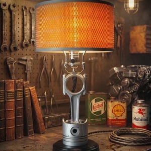 May include: A table lamp featuring a silver metal engine piston and connecting rod as the base, mounted on a circular black stand. The lamp has a cylindrical shade with an orange diamond-patterned mesh exterior and a warm internal light source.