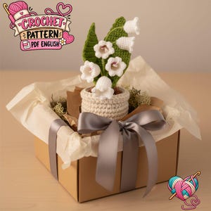 Peut inclure: Une plante muguet au crochet avec des fleurs blanches en forme de cloche et des feuilles vertes dans un pot beige, présentée dans une boîte cadeau en carton brun doublée de papier de soie et nouée d'un ruban satin gris. Texte CROCHET PATTERN PDF ENGLISH.