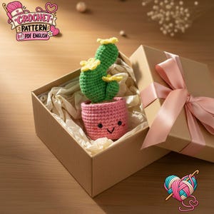 Peut inclure: Un cactus vert crocheté avec des fleurs jaunes dans un pot rose avec un visage souriant, placé dans une boîte cadeau en carton marron ouverte avec un ruban rose clair. Le texte CROCHET PATTERN PDF ENGLISH est visible en haut à gauche.