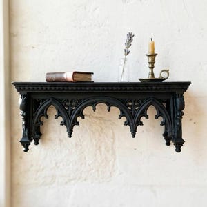 May include: A black wall-mounted shelf featuring intricate pointed arch carvings along the front edge. The shelf surface holds a small brown book, a clear glass bottle containing a sprig of dried lavender, and a brass candle holder with a candle.