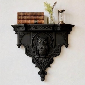 May include: A black, ornate wall shelf with an owl carving. The shelf holds three antique books, a small glass vase with greenery, a feather, and a brass hourglass. The shelf has a decorative, gothic design.