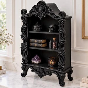 May include: A black, three-tiered shelf featuring ornate, carved scrollwork along the frame and legs. The shelves hold a small lidded container, a skull, two books, a glass bottle with cork, a purple crystal cluster, and a lit candle holder.