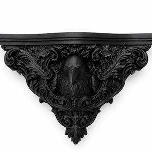 May include: A matte black wall shelf featuring a central sculpted raven head surrounded by intricate, symmetrical acanthus leaf scrollwork. The shelf has a flat top surface and a decorative, pointed bracket base with detailed relief carvings.