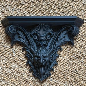 Pode incluir: Prateleira de parede decorativa preta com detalhes intrincados em estilo g&oacute;tico. A prateleira apresenta um rosto diab&oacute;lico com chifres, rodeado por folhas e volutas ornamentadas. A prateleira &eacute; feita de um material s&oacute;lido e tem um topo plano.