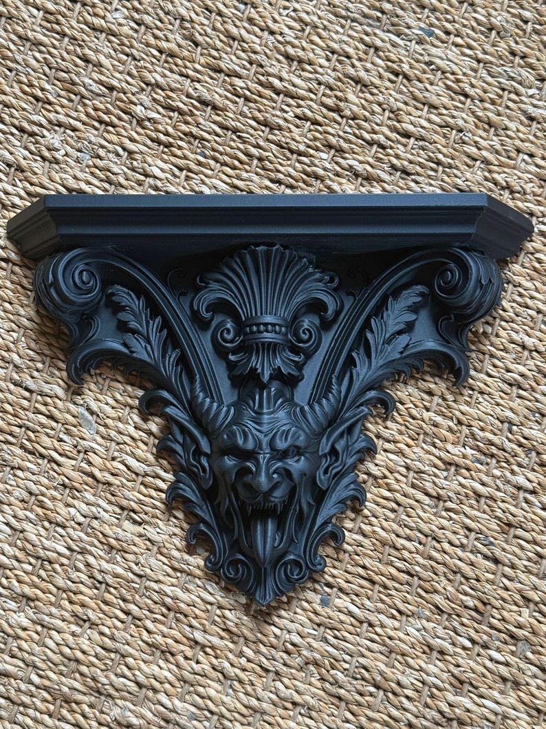 Pode incluir: Prateleira de parede decorativa preta com detalhes intrincados em estilo g&oacute;tico. A prateleira apresenta um rosto diab&oacute;lico com chifres, rodeado por folhas e volutas ornamentadas. A prateleira &eacute; feita de um material s&oacute;lido e tem um topo plano.