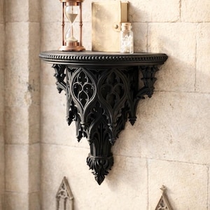 May include: A black, gothic-style wall shelf with intricate carved details. The shelf holds a copper hourglass, a small book, and a glass bottle. The shelf is mounted on a textured, light-colored stone wall.