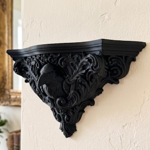 May include: A matte black wall-mounted shelf featuring an intricate, carved design of a raven head surrounded by elaborate scrolling acanthus leaf patterns. The shelf has a flat top surface and a decorative, pointed base extending from the wall.