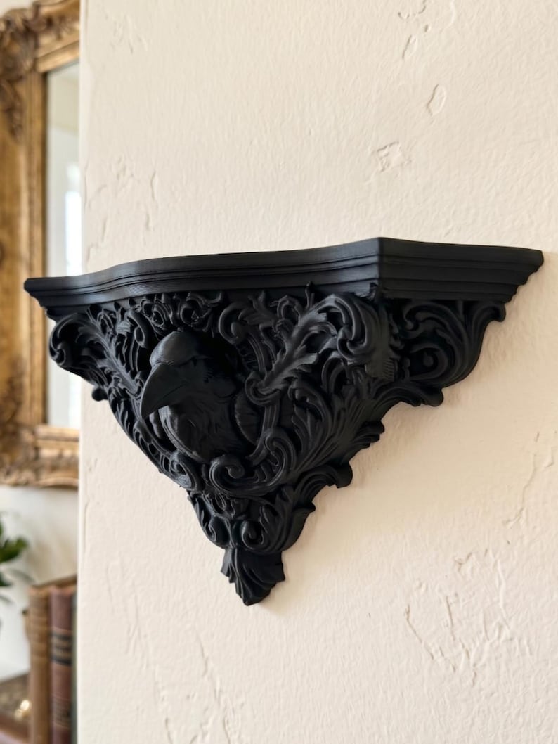 May include: A matte black wall-mounted shelf featuring an intricate, carved design of a raven head surrounded by elaborate scrolling acanthus leaf patterns. The shelf has a flat top surface and a decorative, pointed base extending from the wall.