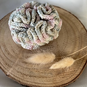 Könnte beinhalten: Ein handgemachtes, mehrfarbiges Häkel-Haargummi in Pastelltönen von Rosa, Grün und Beige. Das Haargummi ist rund und strukturiert und liegt auf einer Holzscheibe mit zwei getrockneten, flauschigen, beigen Hasenschwanzgrasstielen.