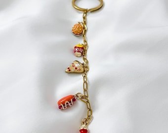 May include: A gold-colored keychain with a ring and chain holding miniature food charms. The charms include a hamburger, a box of fries, a slice of pizza, a hot dog, and a ketchup bottle. The food charms are painted with bright colors.