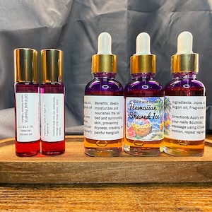 Puede incluir: Una bandeja de madera contiene cuatro botellas de aceite para cut&iacute;culas. Dos peque&ntilde;as botellas roll-on con tapas doradas y etiquetas que dicen "Oil'd and Polish'd. CUTICLE OIL hawaiian shaved ice." Dos botellas m&aacute;s grandes con l&iacute;quido morado, tapas doradas y cuentagotas blancos.