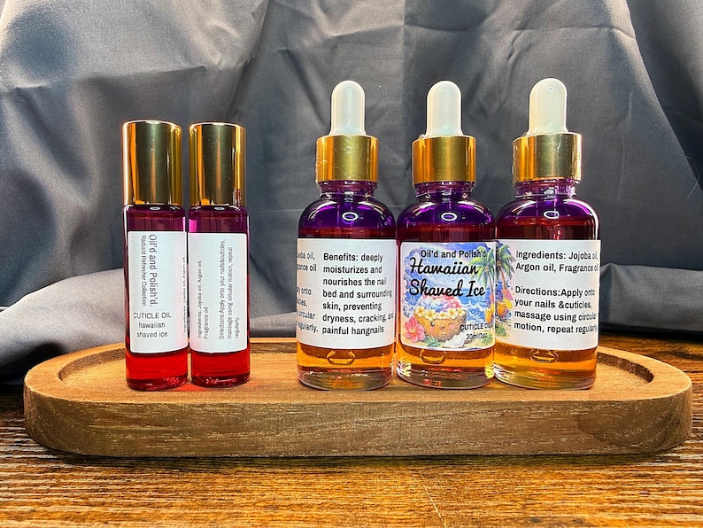 Puede incluir: Una bandeja de madera contiene cuatro botellas de aceite para cut&iacute;culas. Dos peque&ntilde;as botellas roll-on con tapas doradas y etiquetas que dicen "Oil'd and Polish'd. CUTICLE OIL hawaiian shaved ice." Dos botellas m&aacute;s grandes con l&iacute;quido morado, tapas doradas y cuentagotas blancos.
