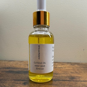 Op de afbeelding: Een heldere glazen flesje nagelriemolie met een gouden dop en witte druppelaar. Het etiket luidt "CUTICLE OIL ocean wave 30ml/1oz". De olie binnenin is geel.