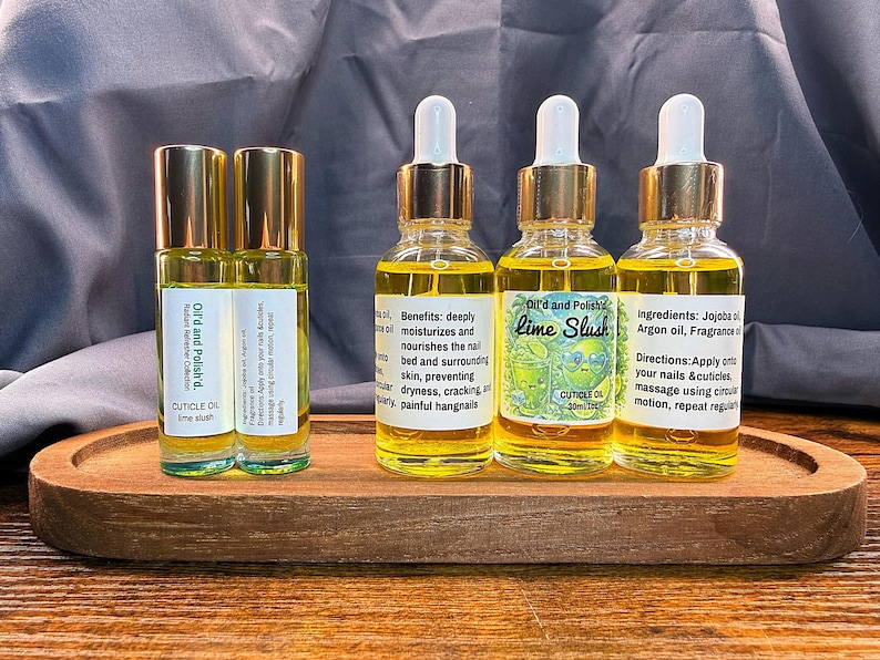 Puede incluir: Una bandeja de madera contiene cuatro botellas de aceite para cut&iacute;culas y dos botellas con rodillo. Las botellas tienen tapas y cuentagotas dorados, llenos de un l&iacute;quido amarillo. Las etiquetas dicen "Lime Slush" y "Cuticle Oil".