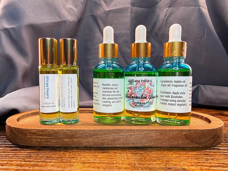 Puede incluir: Una bandeja de madera contiene dos botellas roll-on y tres botellas con cuentagotas de aceite para cut&iacute;culas. Las botellas tienen tapas doradas y etiquetas con texto. Las botellas con cuentagotas contienen un l&iacute;quido azul y verde.