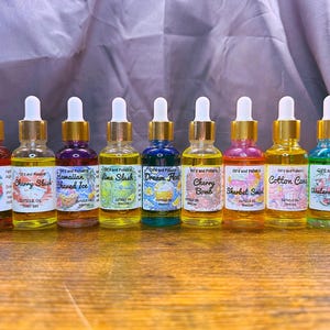 Puede incluir: Una colecci&oacute;n de aceites para cut&iacute;culas en botellas de vidrio transparente con tapas doradas y cuentagotas blancos. Cada botella contiene un aceite de diferente color y tiene una etiqueta con un nombre &uacute;nico, como "Cherry Slush".