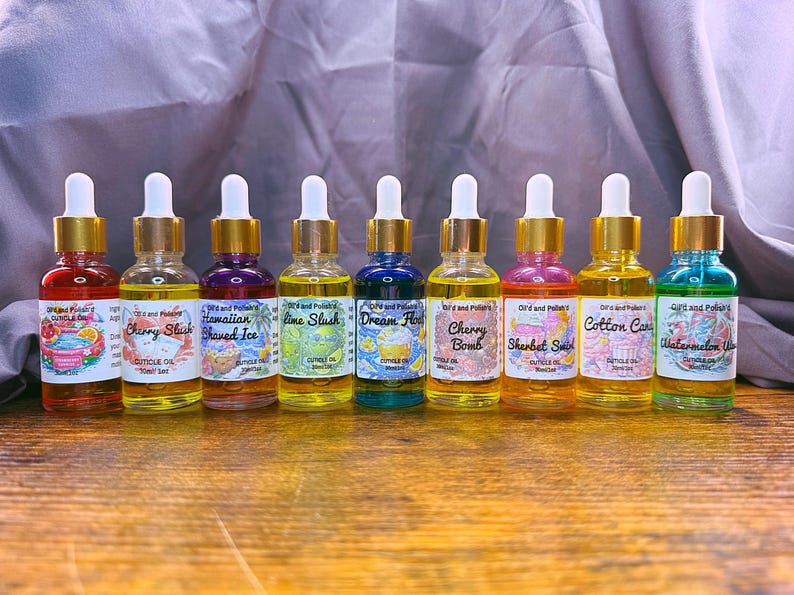 Puede incluir: Una colecci&oacute;n de aceites para cut&iacute;culas en botellas de vidrio transparente con tapas doradas y cuentagotas blancos. Cada botella contiene un aceite de diferente color y tiene una etiqueta con un nombre &uacute;nico, como "Cherry Slush".