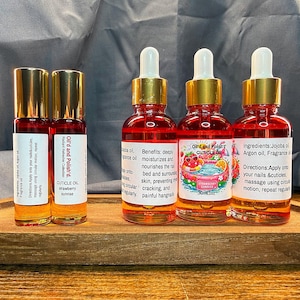 Puede incluir: Una bandeja de madera contiene dos botellas roll-on y cuatro botellas con cuentagotas de aceite para cut&iacute;culas. Las botellas tienen tapas doradas y etiquetas con texto. El aceite es de color rojo-naranja, con "Cuticle Oil".