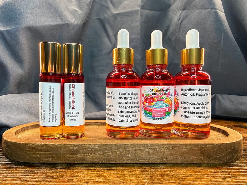 Puede incluir: Una bandeja de madera contiene dos botellas roll-on y cuatro botellas con cuentagotas de aceite para cut&iacute;culas. Las botellas tienen tapas doradas y etiquetas con texto. El aceite es de color rojo-naranja, con "Cuticle Oil".