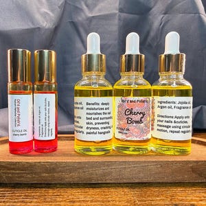 Puede incluir: Una bandeja de madera contiene dos peque&ntilde;os frascos roll-on y tres frascos con cuentagotas de aceite para cut&iacute;culas. Los frascos roll-on tienen tapas doradas y l&iacute;quido rojo. Los frascos con cuentagotas tienen tapas doradas y vidrio transparente con l&iacute;quido amarillo. La etiqueta dice "Cherry Bomb".