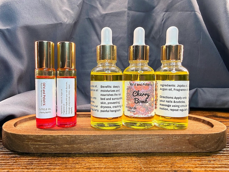Puede incluir: Una bandeja de madera contiene dos peque&ntilde;os frascos roll-on y tres frascos con cuentagotas de aceite para cut&iacute;culas. Los frascos roll-on tienen tapas doradas y l&iacute;quido rojo. Los frascos con cuentagotas tienen tapas doradas y vidrio transparente con l&iacute;quido amarillo. La etiqueta dice "Cherry Bomb".
