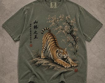 May include: An olive green short-sleeve t-shirt featuring a graphic of a tiger stretching on a rock beneath a flowering tree. The design includes black Chinese calligraphy text and small red square stamps. The shirt has a crew neck and a size L label.