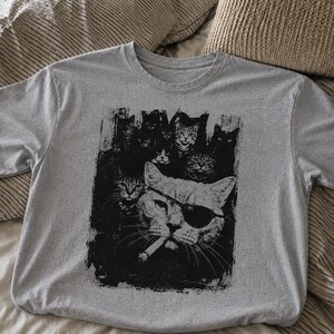 May include: A gray t-shirt featuring a black and white graphic of several cats, including one wearing an eye patch and smoking a cigarette. The cats are arranged in a stylized, artistic design.