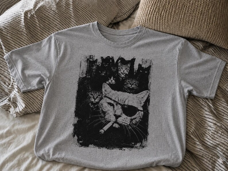 May include: A gray t-shirt featuring a black and white graphic of several cats, including one wearing an eye patch and smoking a cigarette. The cats are arranged in a stylized, artistic design.