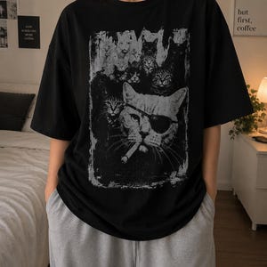 May include: Black t-shirt featuring a distressed graphic of several cats, including one wearing an eye patch and smoking a cigarette. The design is in shades of gray and white. The shirt has short sleeves and a crew neck.