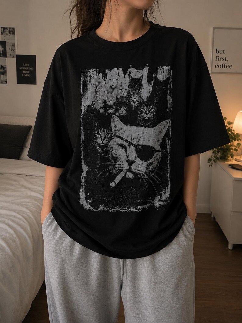 May include: Black t-shirt featuring a distressed graphic of several cats, including one wearing an eye patch and smoking a cigarette. The design is in shades of gray and white. The shirt has short sleeves and a crew neck.