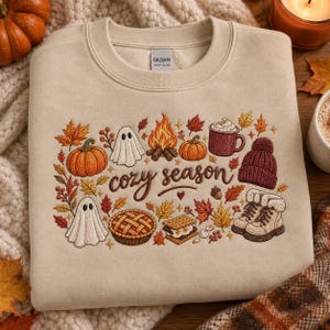 May include: A beige crewneck sweatshirt featuring an embroidered design with the text cozy season. The graphic includes pumpkins, ghosts, a campfire, a mug of cocoa, a knit hat, a pie, a s'more, boots, and various autumn leaves in orange and red.