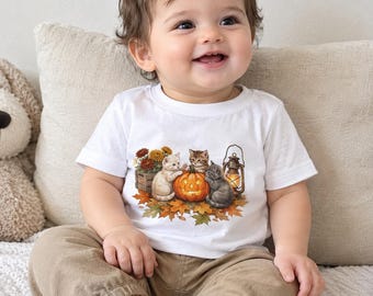 May include: A white short-sleeved t-shirt featuring a graphic print of three kittens sitting around a glowing jack-o'-lantern, a vintage-style lantern, and a wooden crate filled with orange and yellow autumn flowers, all resting on fallen maple leaves.