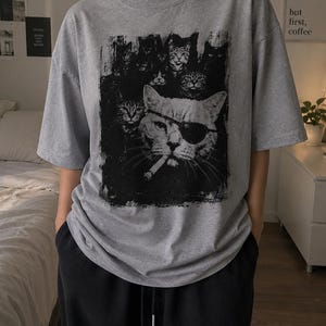 May include: Gray t-shirt featuring a black and white graphic of several cats, with one cat wearing an eye patch and smoking a cigarette. The shirt has short sleeves and a crew neck.