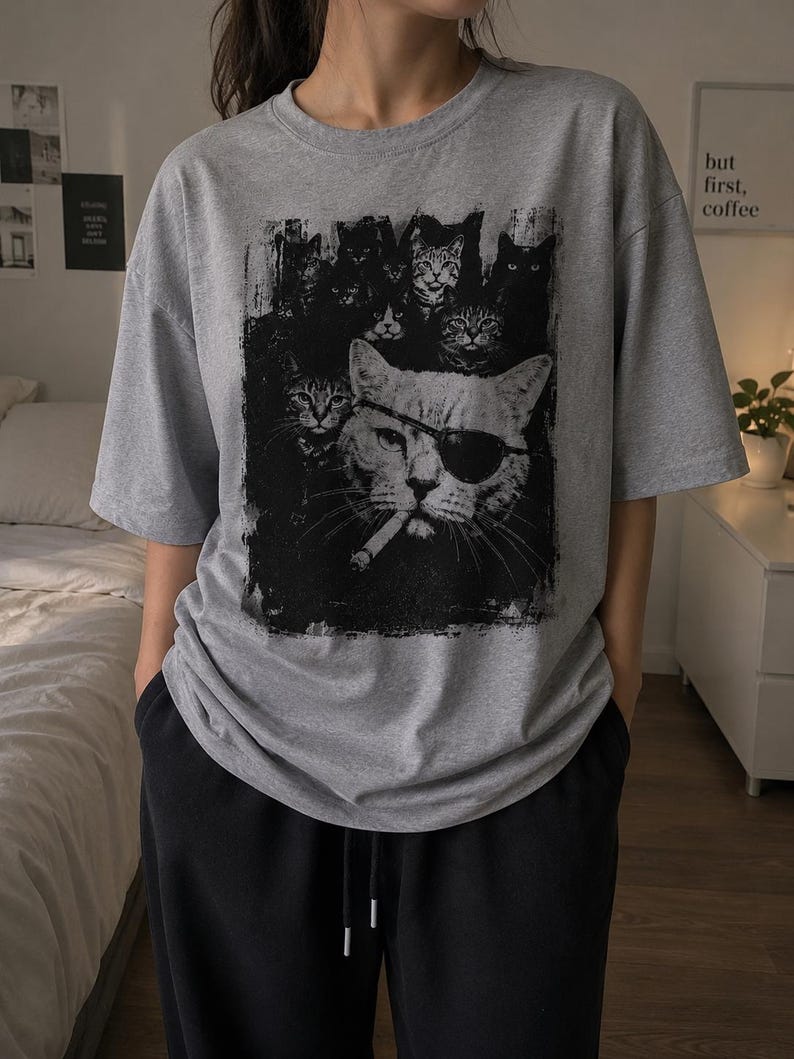 May include: Gray t-shirt featuring a black and white graphic of several cats, with one cat wearing an eye patch and smoking a cigarette. The shirt has short sleeves and a crew neck.