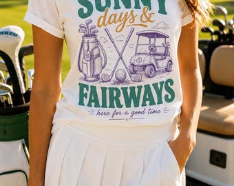 May include: A white short-sleeved t-shirt featuring a graphic print with the text SUNNY days & FAIRWAYS here for a good time. The design includes a golf bag, two crossed golf clubs, a golf ball, and a golf cart in shades of green, gold, and purple.