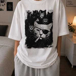 May include: White t-shirt featuring a black and white graphic of several cats, including one wearing an eye patch and smoking a cigarette. The shirt has a crew neck and short sleeves. The text "but first, coffee" is visible in the background.