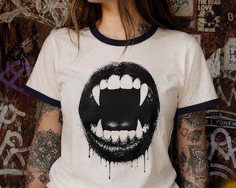 May include: A cream-colored ringer t-shirt with dark navy blue trim on the collar and sleeves. The center of the shirt features a large, black graphic print of an open mouth with prominent vampire fangs and dripping ink-like details.