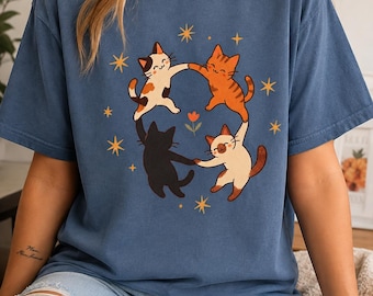 May include: A blue short-sleeve t-shirt featuring a graphic print of four cats in different colors—calico, orange tabby, black, and Siamese—holding paws in a circle around a small orange flower, surrounded by several yellow four-pointed stars.