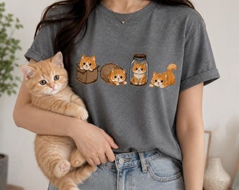 May include: A charcoal gray short-sleeve t-shirt featuring a graphic print of four orange tabby kittens in various positions, including one inside a brown cardboard box, one in a brown sack, one inside a glass jar, and one lying down.