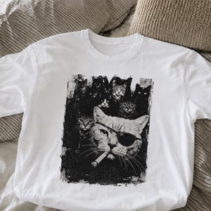 May include: White t-shirt featuring a black and white graphic of several cats. The central cat wears an eye patch and sunglasses, with a cigarette in its mouth. Other cats are in the background, creating a unique design.