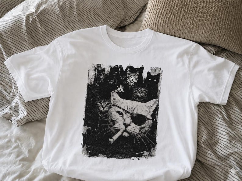 May include: White t-shirt featuring a black and white graphic of several cats. The central cat wears an eye patch and sunglasses, with a cigarette in its mouth. Other cats are in the background, creating a unique design.