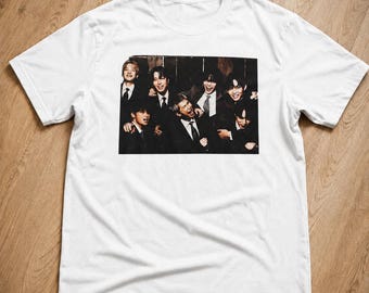 May include: A white short-sleeved t-shirt featuring a rectangular graphic print on the chest. The print displays a group of seven individuals wearing dark suits, white shirts, and ties, captured in a candid, smiling pose against a dark background.
