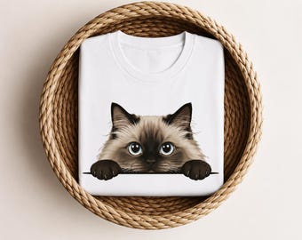 May include: A white t-shirt featuring a graphic illustration of a fluffy Siamese cat with large blue eyes and dark brown paws peeking over a horizontal line. The cat has a light tan face with a dark brown mask and pointed ears.
