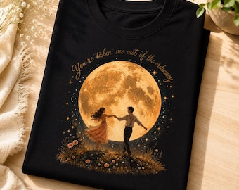 May include: A black t-shirt featuring a graphic of a couple dancing in a field under a large, glowing yellow moon. The text "You're takin' me out of the ordinary" is printed in a cursive font above the moon, surrounded by small stars.