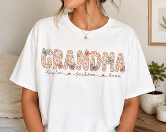 May include: White t-shirt with the word "GRANDMA" in a floral design, featuring butterflies. Below the word are the names "taylor", "jackson", and "emma". A casual, comfortable tee.