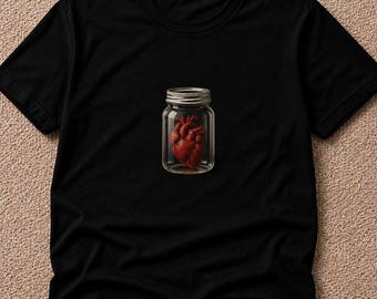 May include: A black short-sleeved t-shirt featuring a graphic print in the center. The design depicts a realistic red anatomical human heart enclosed within a clear glass mason jar with a metallic screw-top lid, set against the solid black fabric.